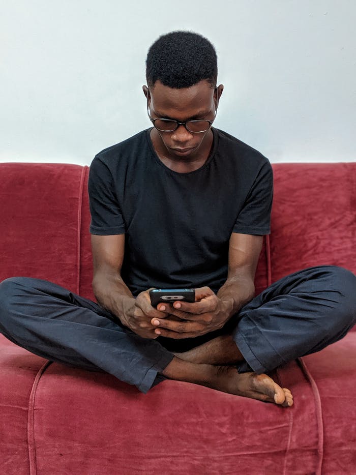 African man sitting on a red sofa indoors, using a smartphone, focused and relaxed.