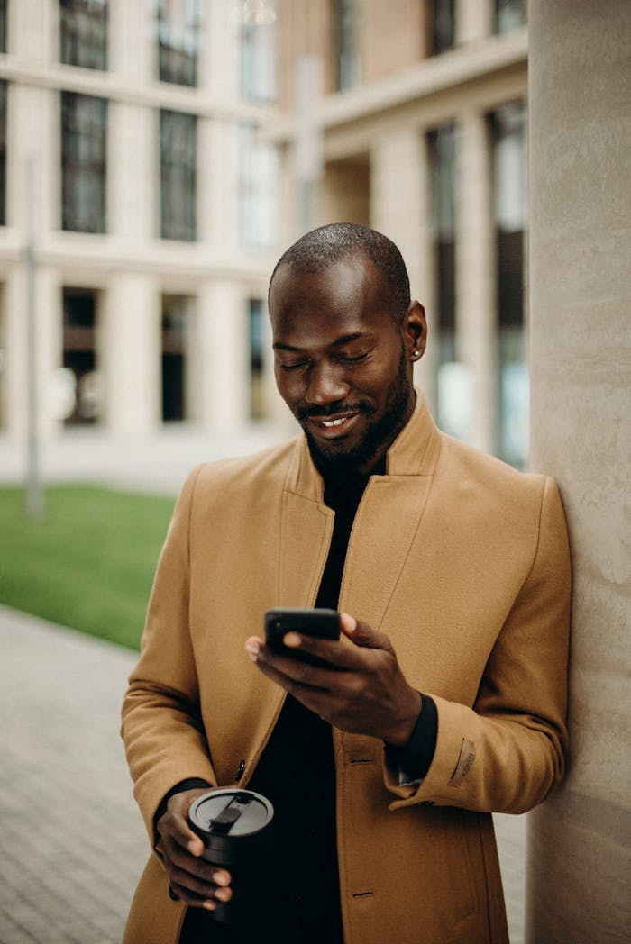 Relaxed man in coat smiles while using smartphone and holding coffee outside.