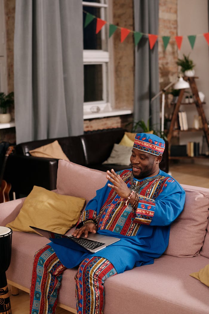 Happy man in dashiki, using a laptop for video call, sitting on a cozy couch indoors.