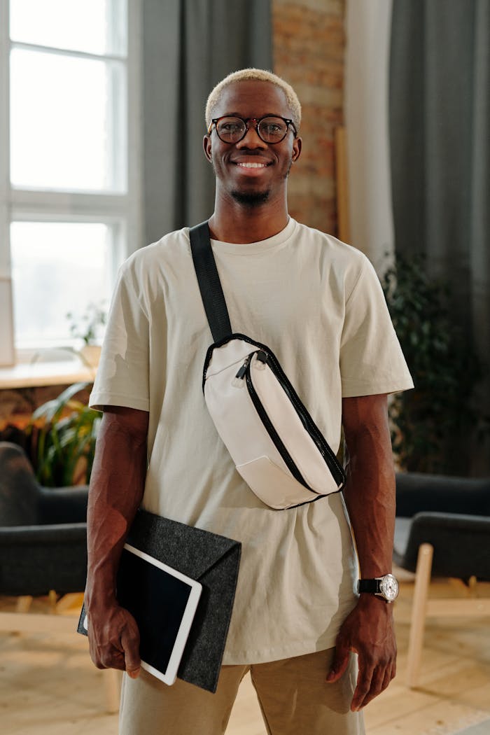 Smiling young man holding a tablet and wearing a belt bag indoors, exuding confidence and style.