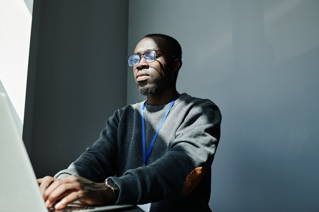 Black man wearing eyeglasses and gray sweater typing on laptop in a sunlit room.